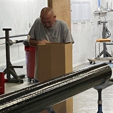 A man in a grey tshirt is working in a workshop setting with his head down. The background features some heavy machinery of some kind.