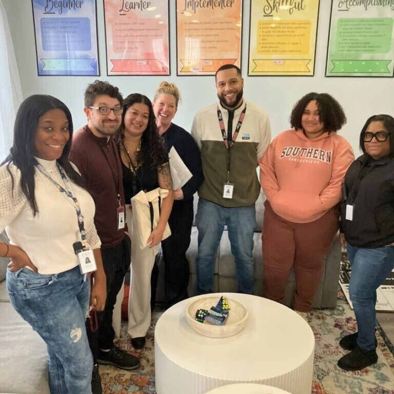 Image shows 7 individual staff members of Lisa's House posing together within the program office. 2 of the staff members are male and 5 of them are female. There are 5 blue, red, orange, yellow, and green photo frames in the background.