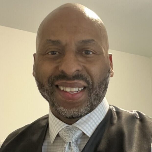Image shows a portrait photo of a man in formal attire (with his grey tie visible) smiling at the camera.
